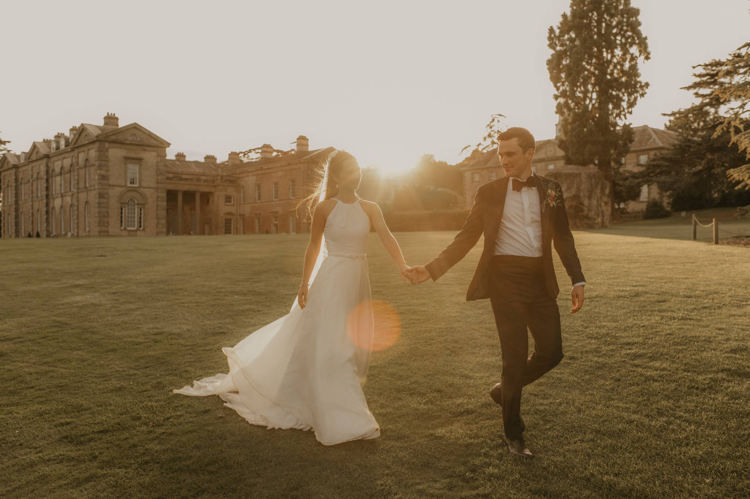 A bride and groom walk across a lawn in the golden light with a neoclassical building in the background