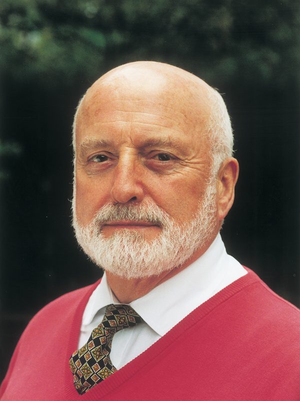 This image shows a white man with a white beard and no hair wearing a red jumper, shirt, and tie. This is Sir Peter Moores
