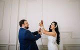 The image shows a joyful newlywed couple pulling on a sally. The bride, wearing a white lace wedding gown with a floral hair accessory, is laughing with her arms outstretched pulling on the rope. The groom, dressed in a navy blue suit, is gazing at his bride affectionately while holding his hands to the same rope. The scene captures a candid and blissful moment between the couple against a plain white wall background in the chapel.