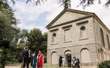 The image shows a wedding party standing outside an old stone chapel with classical architectural features. The building has a triangular pediment roof and arched doorways and windows. In the foreground, there is a group of formally dressed people gathered, with men in suits and women in colourful dresses or saris. They appear to be posing for photographs or celebrating together near the historic building, which is surrounded by trees and landscaped gardens.