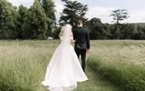 A bride in a long flowy white dress and veil with a groom in a black suit walk away from us on a path into a bright green meadow with large trees and tall grass.