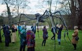 A group of people are gathered around a large, abstract metal sculpture of a spider-like creature in an outdoor park setting. Some are taking photographs of the sculpture, while others observe it from different angles, appreciating the artistic installation.