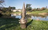 A tall, twisted bronze sculpture resembling a pine cone or flame shape stands near the edge of a pond, reflected in the still waters, with a grand stone house visible in the background surrounded by trees.