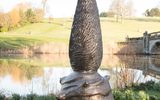 A tall, twisted bronze sculpture resembling a pine cone or flame shape stands near the edge of a pond, reflected in the still waters, with a bridge in the background.