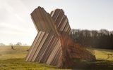 A massive curved wooden structure made of parallel slats stands in an open grassy field against a cloudy sky.