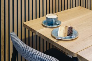 A cake and coffee on a wooden table with a black and white fabric chair and wood panelled walls.