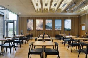 Rows of wooden tables with black and white fabric chairs make up a cafe with light up artworks of a volcano on wood panelled walls.