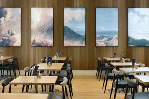 Rows of wooden tables with black and white fabric chairs make up a cafe with light up artworks of a volcano on wood panelled walls.