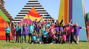 This image shows a group of children in blue, purple and pink tshirts lifting their arms up in celebration posing for a group photo in front of a colourful sculpture outdoors.
