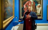 This images shows a woman drinking out of a champagne flute smiling and holding what appears to be an interactive part of an art installation. She is stood in a bright blue art gallery with grand paintings of landscapes and portraits on the wall with gold frames.