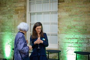 Two women, one older one younger, talk to each other in smart attire and name badges which suggests a work function, against a historic stone wall lit up with green lights and a grand window.