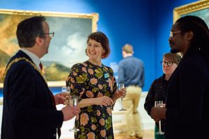 A group of people in smart attire stand and talk with champagne flutes in a blue art gallery that has beautiful landscape paintings in gold frames on the wall.