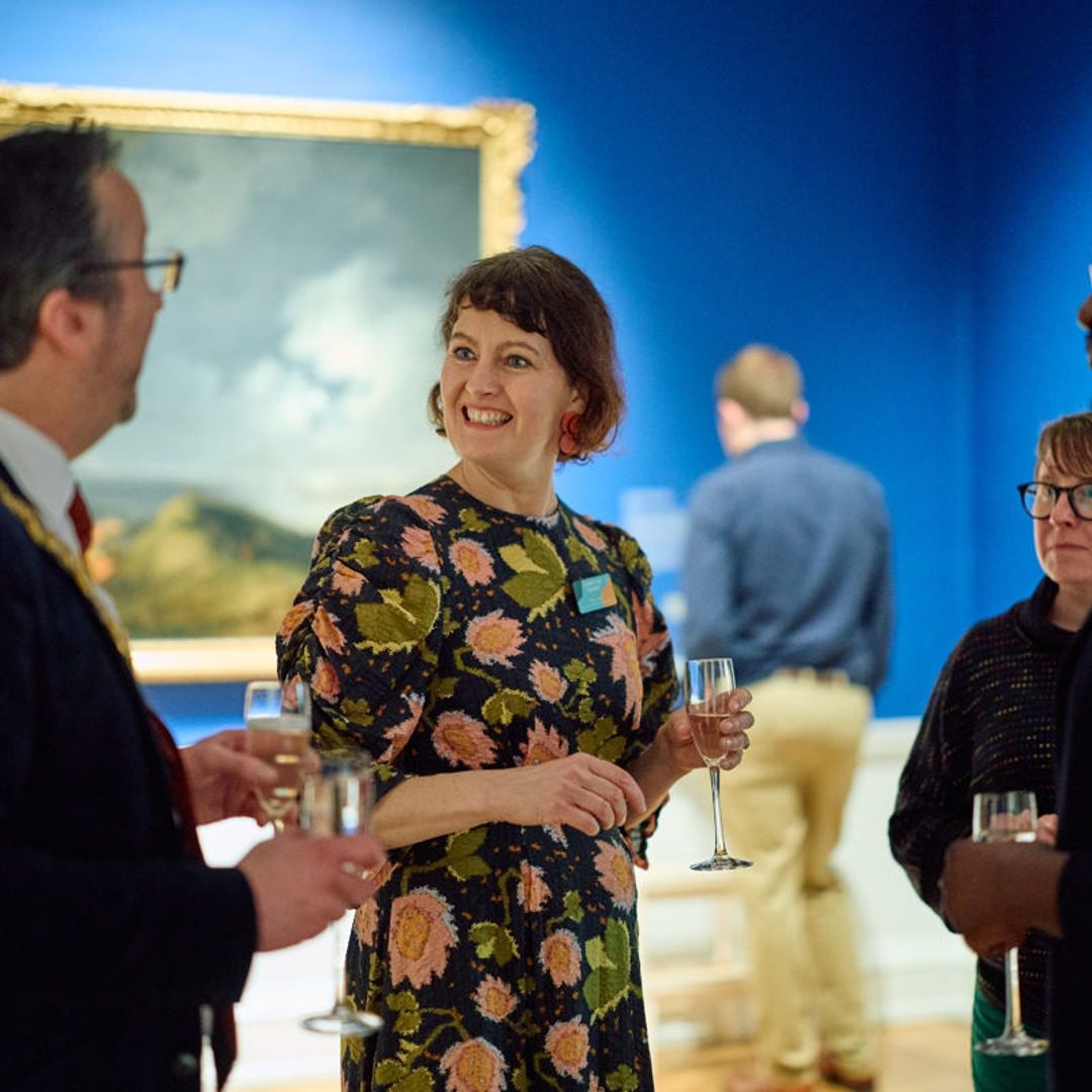 A woman wearing a colourful floral dress is holding a wine glass and smiling at a social gathering or networking event. There is a framed painting behind her and other people visible in the background, also holding drinks.