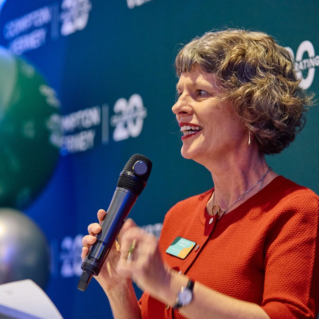 A woman with curly grey hair is speaking into a microphone at a public event. She is wearing a red dress and has a name badge clipped to it. The background shows signage and branding for the event.