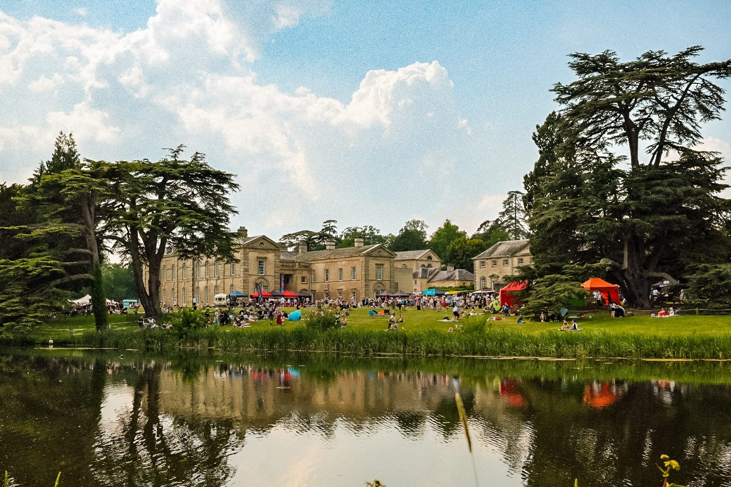 A lake is in the foreground with Compton Verney House the main focus. There are people and food stalls outside the house enjoying food and drink on a summers day.