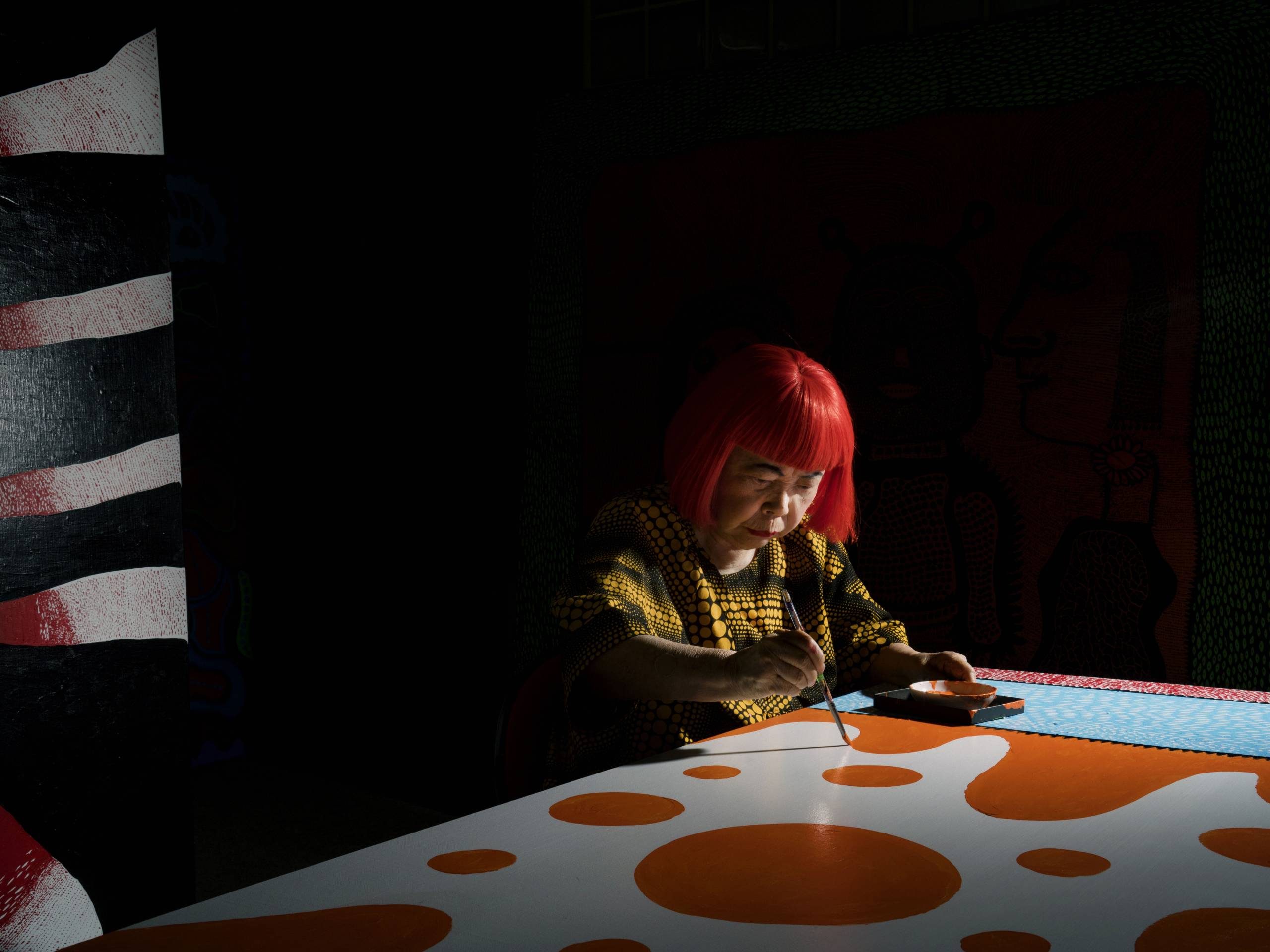 A woman with bright red hair, wearing a yellow and black patterned shirt, hunched over a round, polka-dotted table or surface. She appears to be working on something, and the background is completely black.