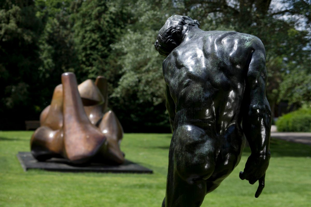 Two large bronze sculptures in an outdoor park. The larger sculpture depicts a muscular male figure in a dynamic, twisting pose. The smaller sculpture in the foreground appears to be an abstract form with smooth, undulating shapes.