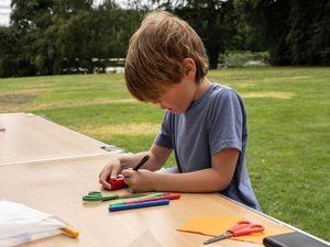 A boy sits outside at a table wearing a grey t-shirt and felt tip pens.