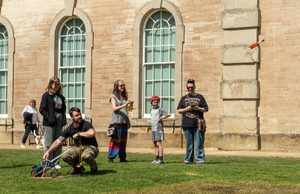 A group of adults and children stand on the lawn with a stone coloured mansion in the background, looking in the distance,