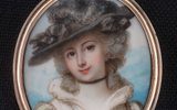 A close-up image of an oval-shaped miniature portrait painting encased in an ornate golden frame. The portrait depicts a woman wearing an ornate hat, with her curly hair styled in an 18th or 19th-century fashion.