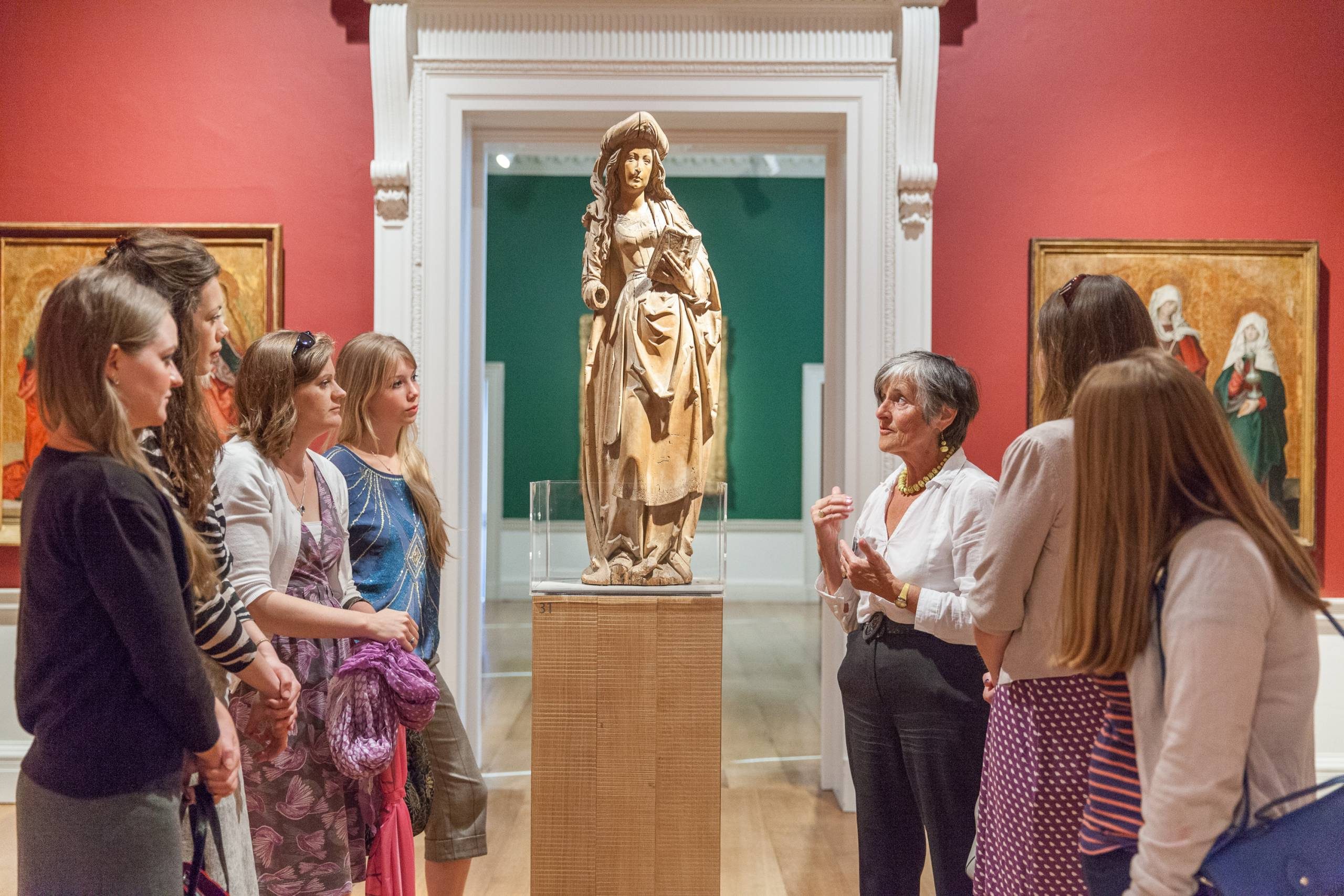 A group of people gather round an artwork on a plinth in a room with red walls and a white archway in the background