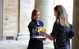 This image shows a woman with a tray of champagne and other drinks offering it to another woman who takes a glass. They are stood amongst large classical columns on what looks like a historic site.