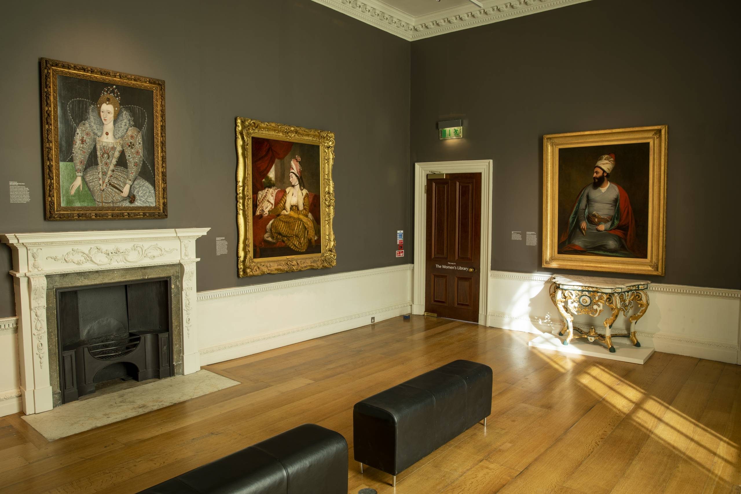 This image depicts an art gallery displaying various artistic works from different eras and cultures. The room features ornate wooden panelling, a decorative fireplace, and large framed paintings hung on the walls, some in gilded frames. One painting shows a regal figure, likely a monarch or nobility, in an elaborate dress and ruff collar. Another portrays an individual in traditional Middle Eastern attire and a turban. In the center of the room is a carved wooden bench or seating area, as well as an ornamental sculptural piece, possibly an antique table or stand with intricate designs. There is also a modern exit sign and a door leading to another part of the gallery.