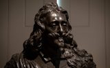 This image shows a close up shot of bronze bust of King Charles I in front of closed shutters that light barely peaks through.
