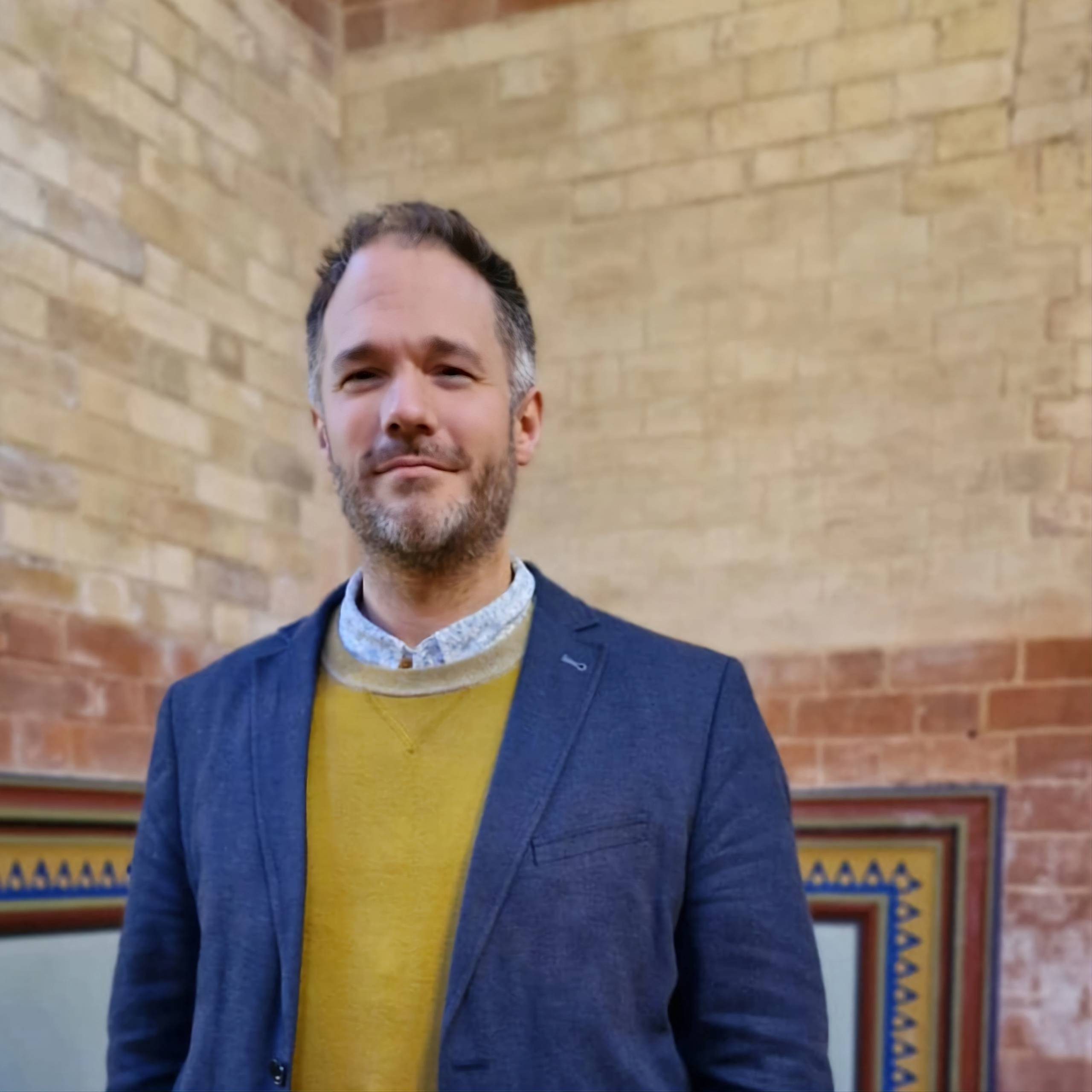 A man with a beard is standing in front of a brick wall. He is wearing a navy blue suit jacket, a yellow sweater, and a patterned shirt. He has a friendly expression on his face.