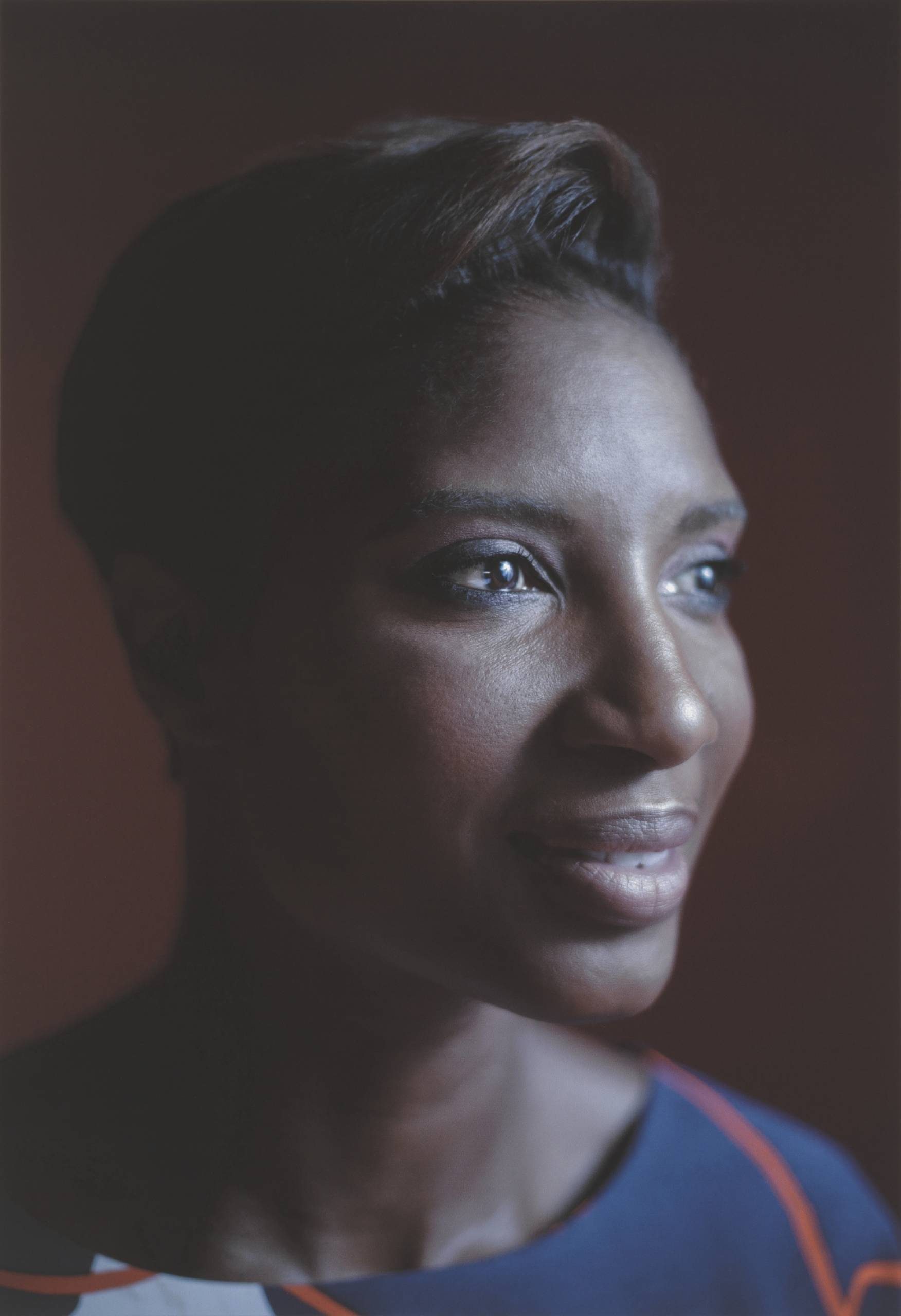 A close-up portrait photograph of a Black woman with brown eyes, smooth skin and short brown hair, her head slightly turned to one side against a dark background, wearing a blue shirt with orange details.