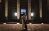 A romantic newlywed couple shares an intimate embrace, with the bride in an elegant gown and the groom in a suit, silhouetted against large arched windows as the evening light casts a warm glow around them in this stunning wedding portrait.