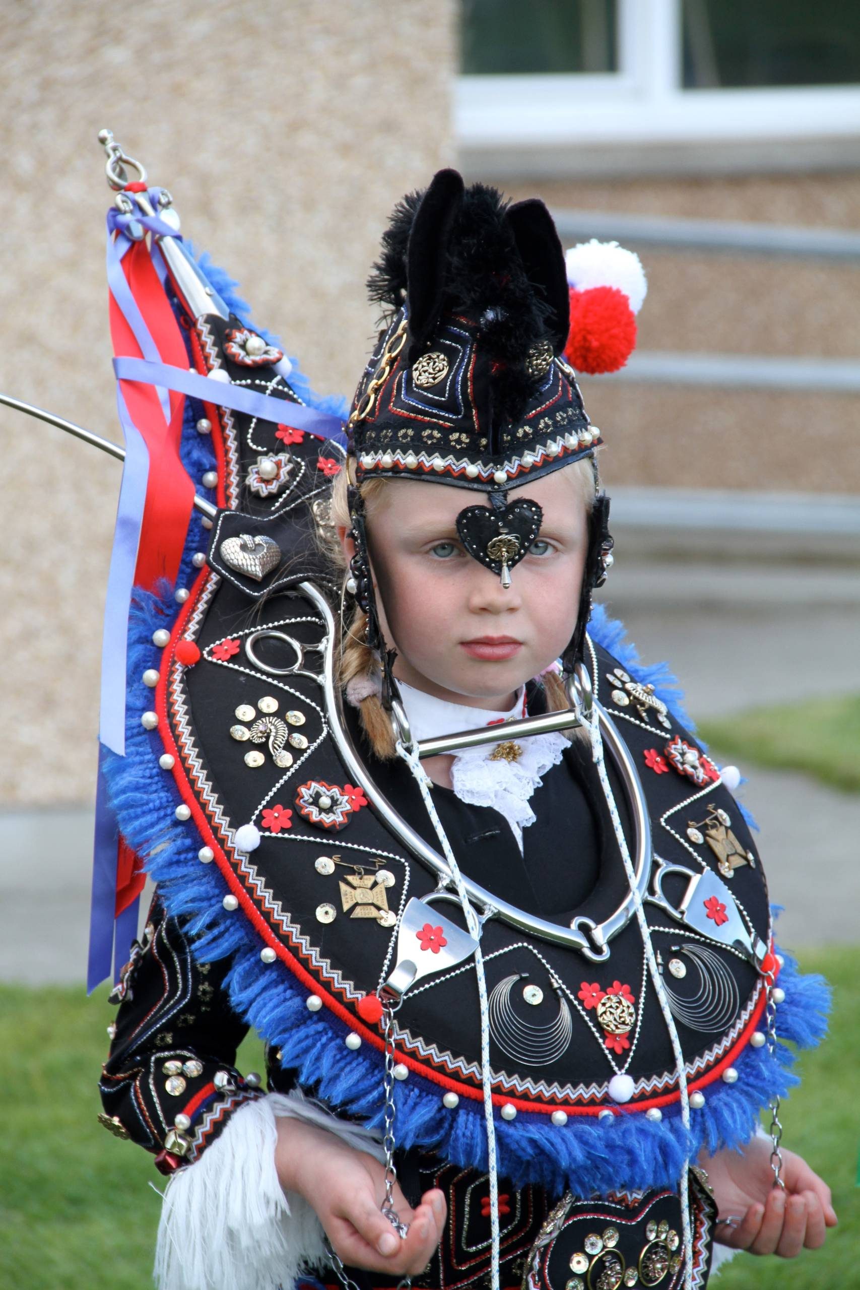 A young girl dressed in an elaborate and heavily embellished traditional cultural costume, carrying a ceremonial staff or flag, posing outdoors against a building backdrop.