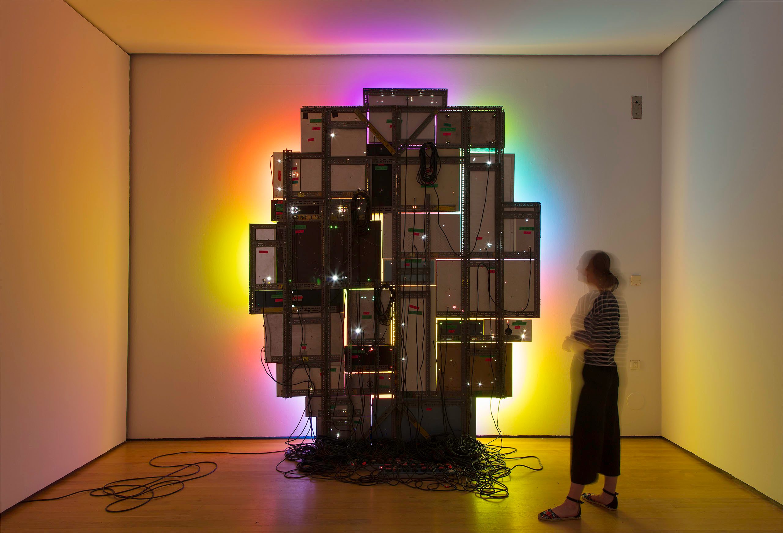 A room installation with a large sculptural structure made of computer monitors, cables, and electronic components, emitting colourful lights and creating an immersive glowing ambiance. A person stands observing the artwork.