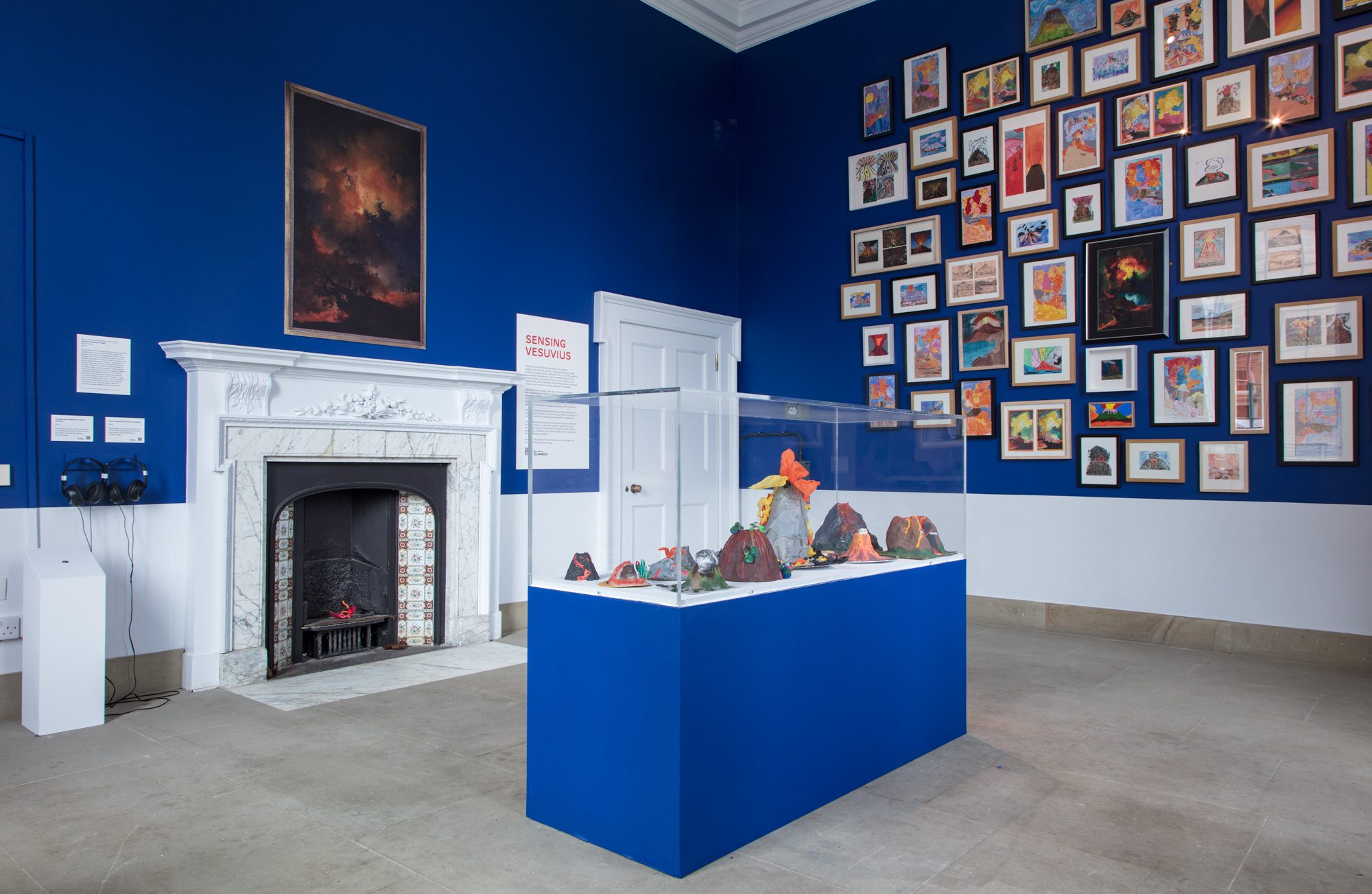 An art gallery exhibition space with deep blue walls displaying numerous framed artworks, featuring a large landscape painting, a decorative fireplace, information panels, and a central pedestal with sculpted forms exhibited.