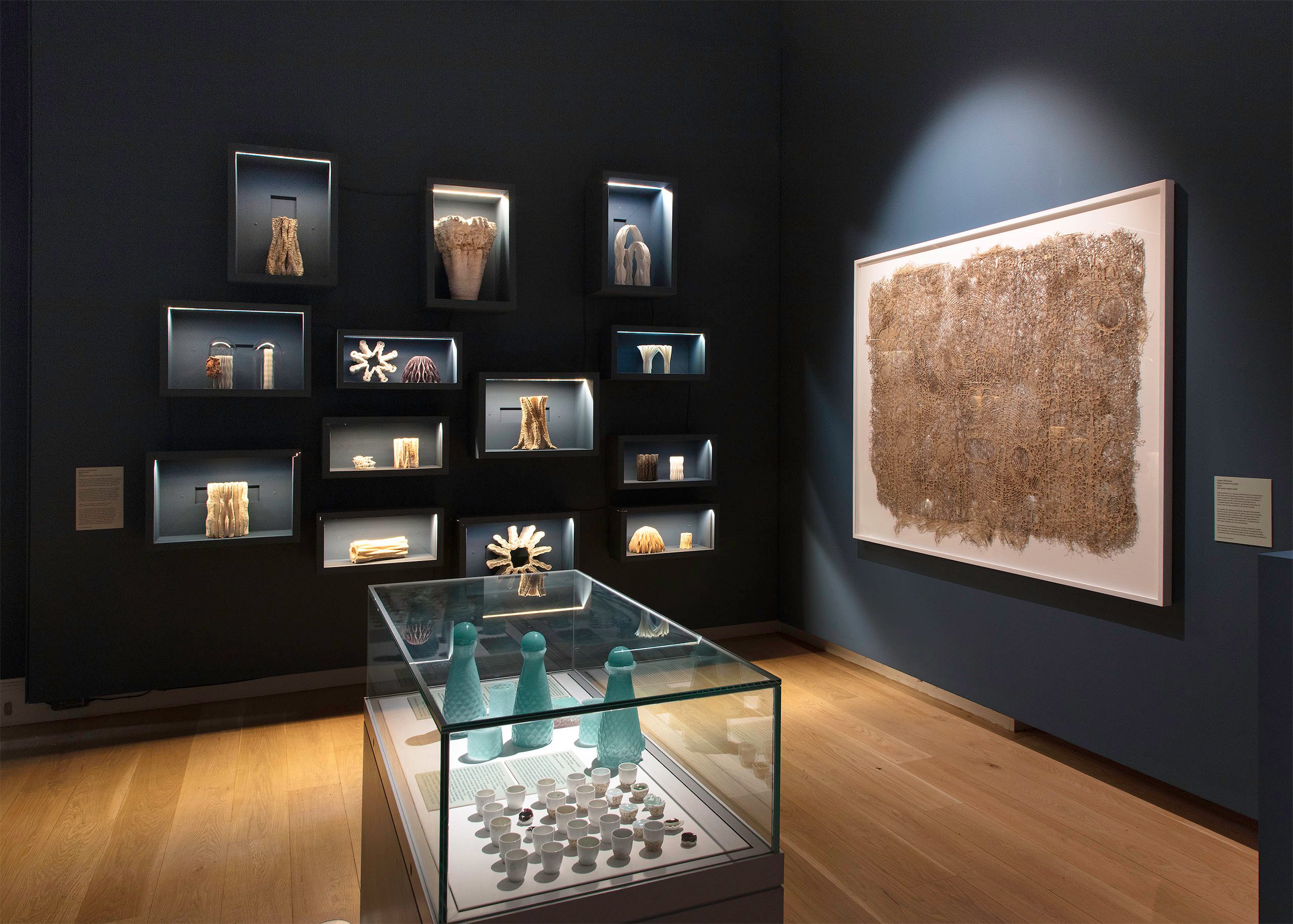 A museum exhibition showcasing various archaeological artifacts and textile pieces displayed in illuminated cases and on the walls. The exhibits appear to be ancient relics, textile fragments, and ceramics, presented in a dimly lit gallery space.