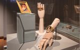 A wooden artist's hand model emerging from behind a curtain. The hand is holding a black ribbon or cord with another portrait miniature pendant attached.