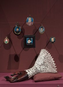 Installation view, The Reflected Self. © Compton Verney, photo by Jamie Woodley (35) Several small portrait miniatures are shown here, and other historical objects. There's a wooden prosthetic hand with intricate lace cuff visible, along with various framed miniature portraits arranged on a burgundy background.