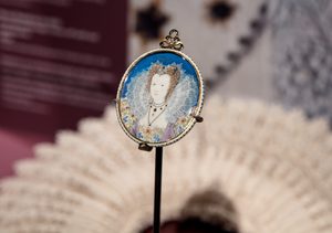 Installation view, The Reflected Self. © Compton Verney, photo by Jamie Woodley (45) A close-up of a single miniature portrait, showing a woman with an ornate lace collar and headdress against a vivid blue background. The portrait is framed in an ornate gold setting.