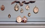 a collection of oval portrait miniatures hanging on a wall. These small, detailed paintings depict various individuals, mostly women, in period dress and hairstyles. A brown ribbon is draped above them.