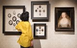 A museum visitor in a yellow shirt examining a wall display of framed portrait miniatures and a larger portrait painting. The miniatures are arranged in groups within their frames.