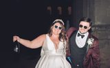 A joyful bride wearing a white sleeveless gown and heart-shaped sunglasses laughs with her groom, who is wearing a burgundy suit and bowtie, as they hold a bottle of celebratory champagne at their wedding reception.