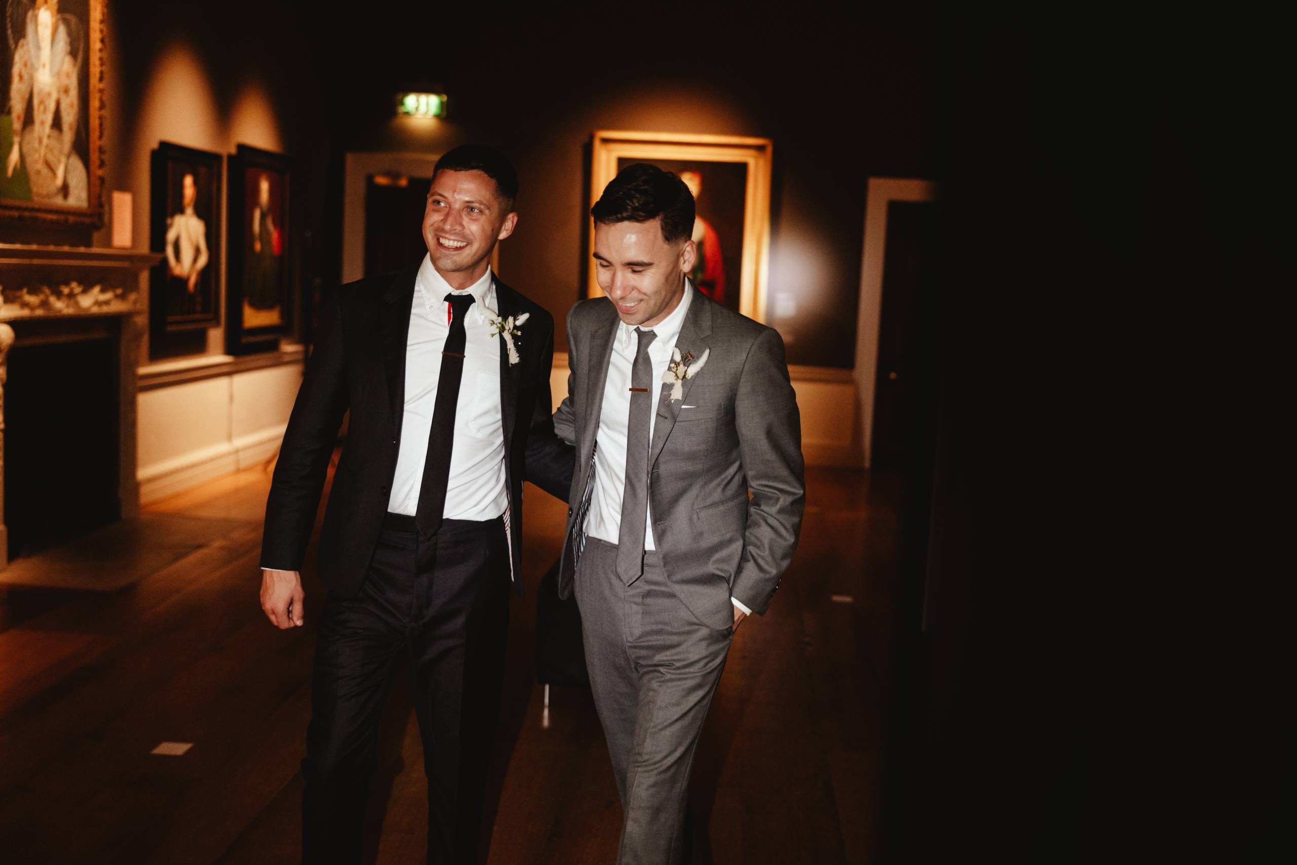 The image shows two grooms, smiling and standing together in what appears to be an art gallery. One man is wearing a black suit with a white shirt and black tie, while the other is wearing a grey suit. They both have boutonniere flowers pinned to their lapels, suggesting this is from their wedding celebration. The warm lighting and framed artwork on the walls creates an elegant, intimate ambiance for this joyful moment between the two grooms or partners.