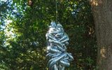 A metallic silver sculpture hanging from a tree branch in a forest setting. The sculpture has an abstract, twisted form resembling a human figure. It's suspended by a wire against a background of green foliage.