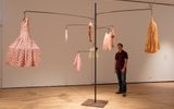 An art installation featuring clothing and fabric items hanging from a metal structure. Various dresses, undergarments and fabric pieces in pink, yellow and flesh tones are suspended from curved metal arms. A person is standing to the right, observing the installation.