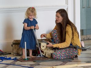 A small girl stands looking at a pole in her hand, with an adult engaging with the child, wearing a yellow cardigan in a room with white walls.