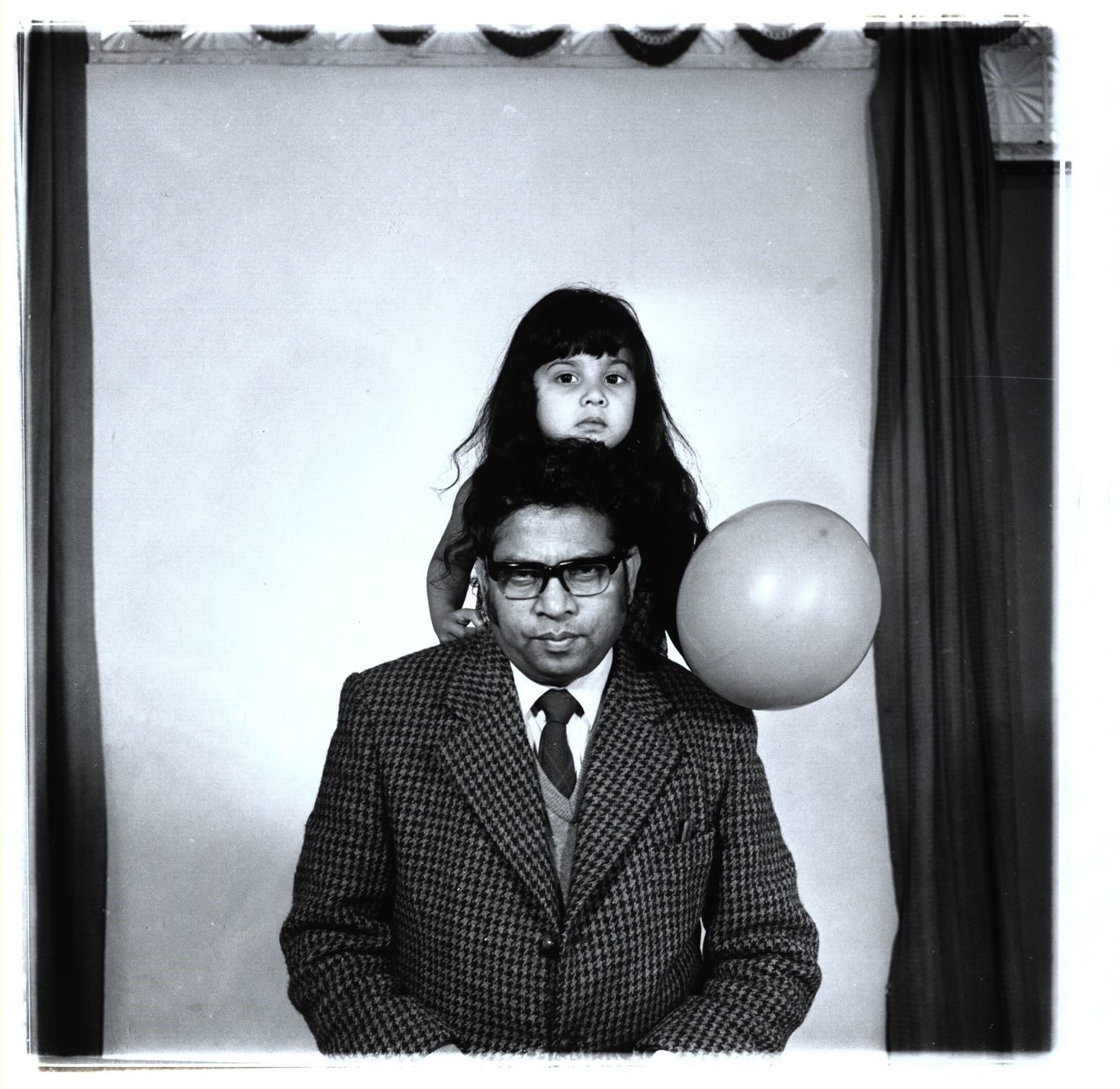 A surreal portrait of a man in a suit and tie with a young girl perched on his shoulders, holding a large white balloon.