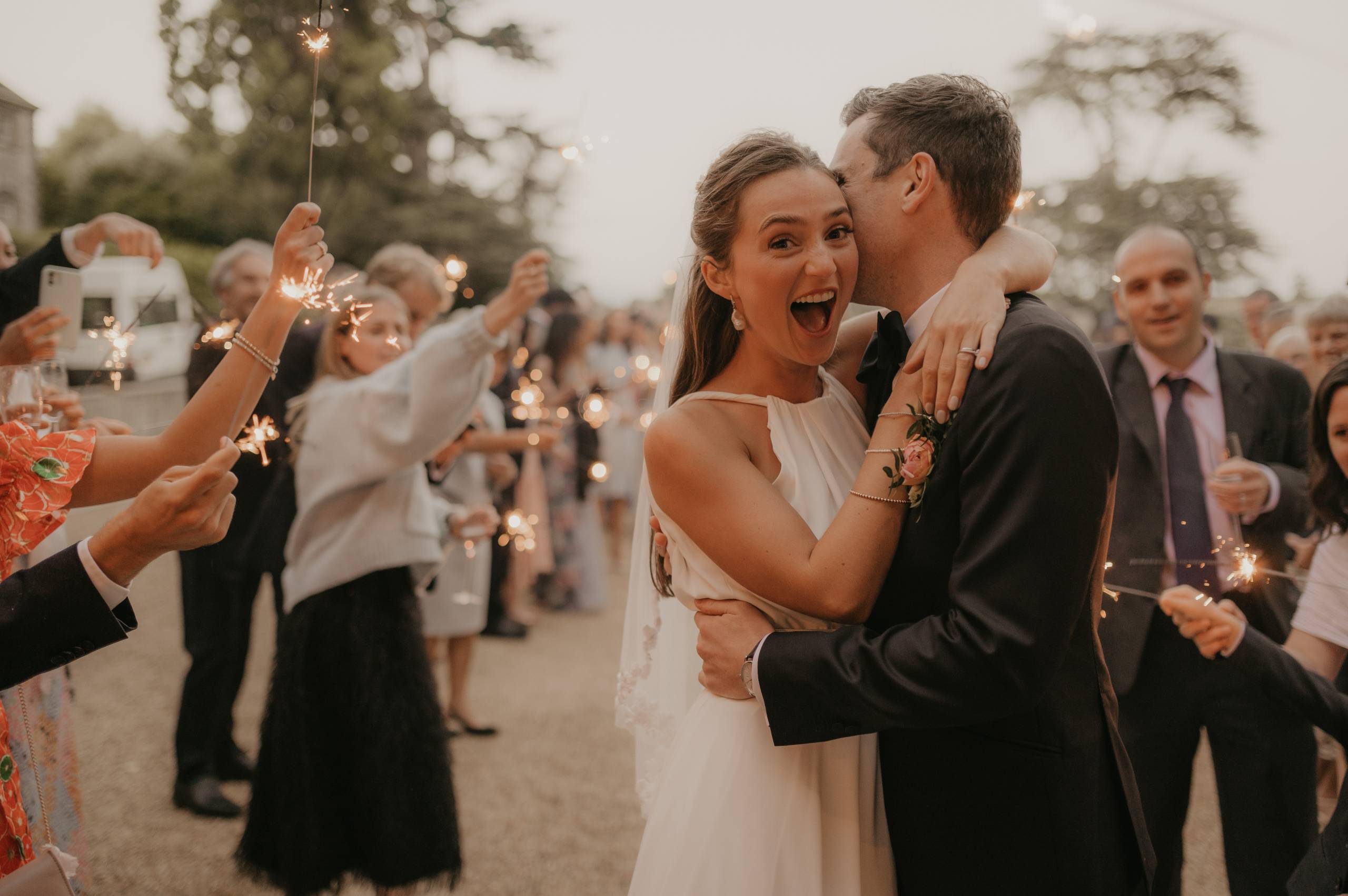 The image shows a joyful newlywed couple at their wedding reception. They are standing outdoors surrounded by trees and string lights, with a crowd of guests in the background. The bride is wearing a sleeveless white wedding gown with a simple belt detail. She has her hair styled up. The groom is wearing a classic black tuxedo with a bow tie. Both have huge smiles on their faces as they celebrate this special moment together amidst the festive reception setting. The lighting creates a warm, romantic ambiance capturing their happiness and excitement on their wedding day.