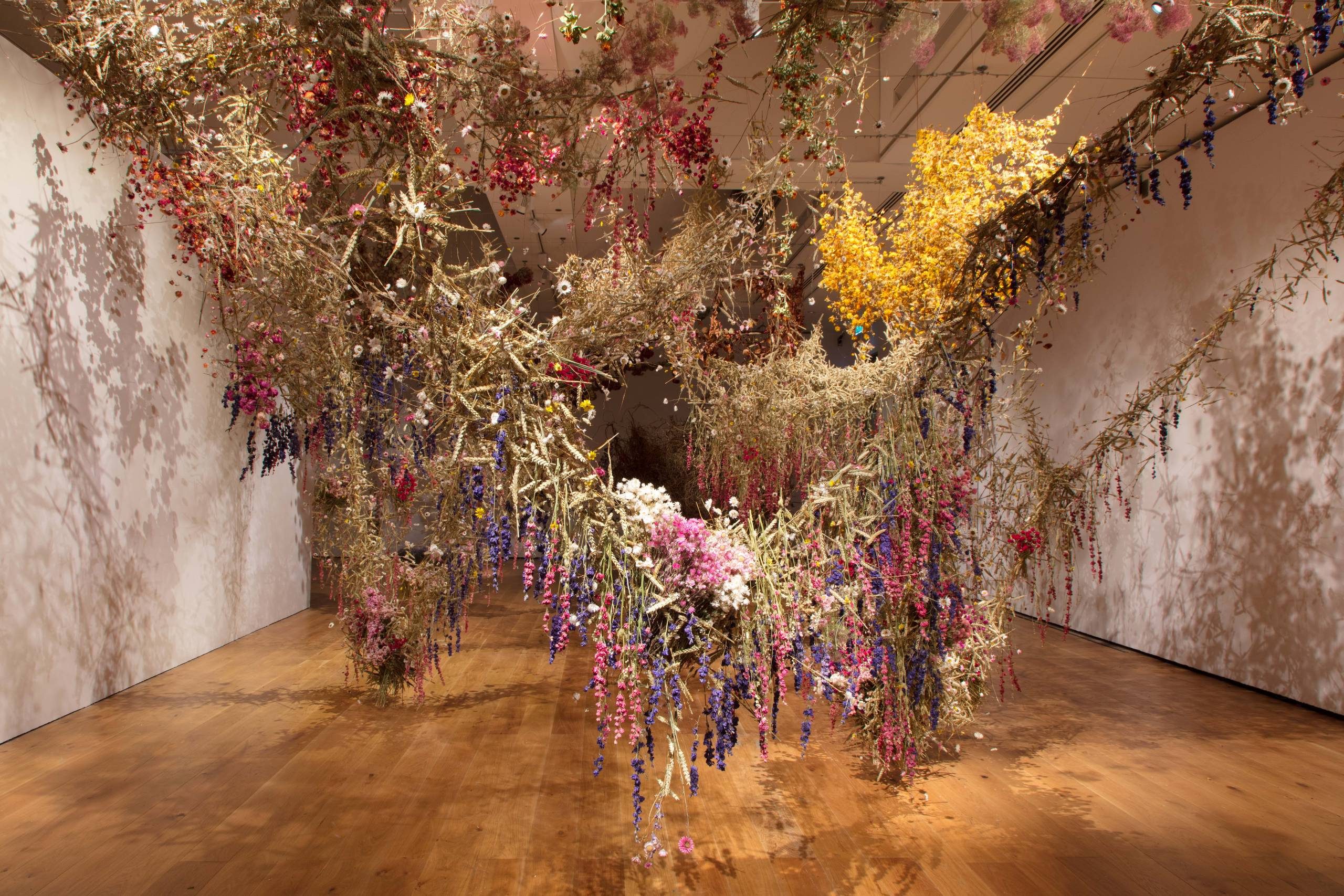 An immersive art installation featuring a room filled with hanging dried flower arrangements and plant materials in rich colours like yellow, red, purple, and white, creating an intricate canopy.
