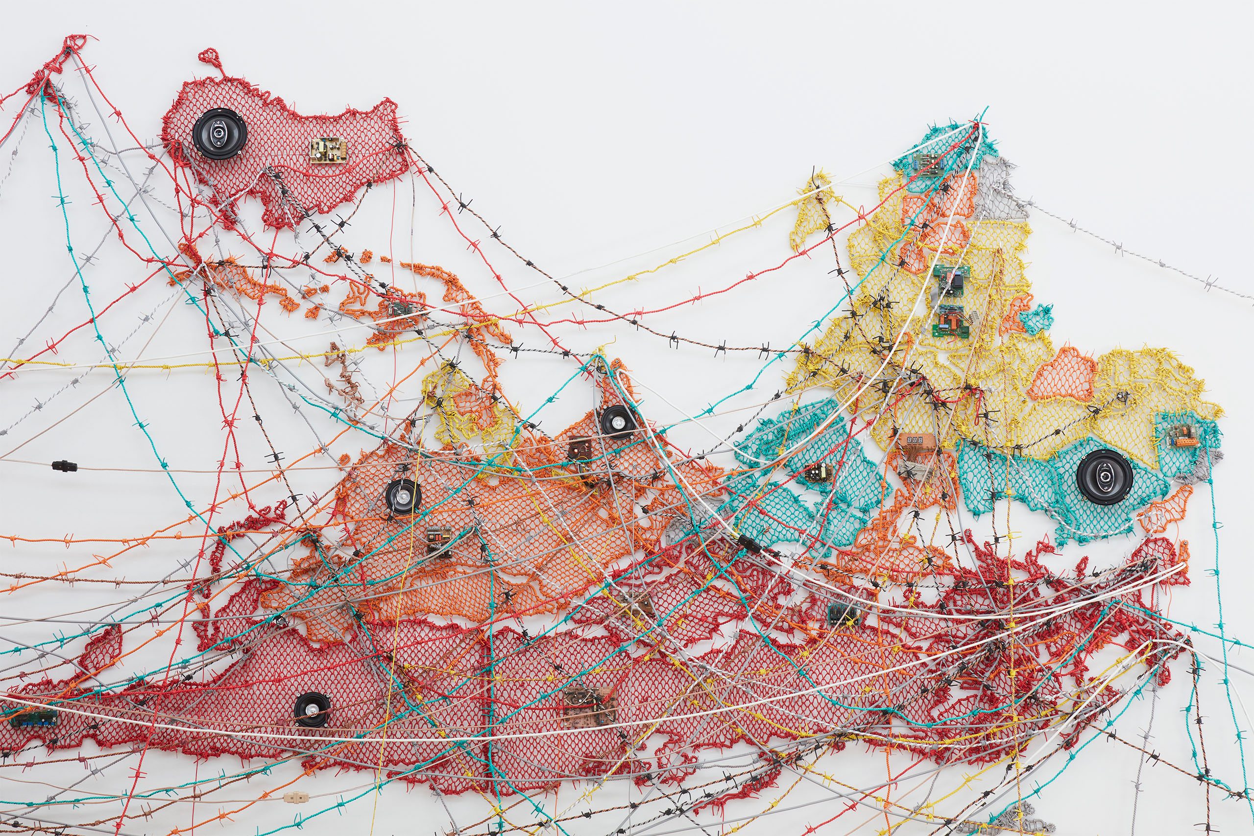 An abstract mixed-media artwork featuring an intricate web-like structure made of mesh, wires, and electronic components in vibrant shades of red, yellow, and turquoise. The piece incorporates various textures and materials, creating a complex and dynamic visual composition.