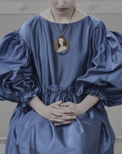 The Reflected Self. © Simon Bevan This image shows a close-up of a person wearing a blue Renaissance-style dress with puffed sleeves. The person's hands are clasped at the waist. Around their neck is a chain with a pendant featuring a miniature portrait of a woman with curled hair wearing a blue dress. The background appears to be a pale wall.